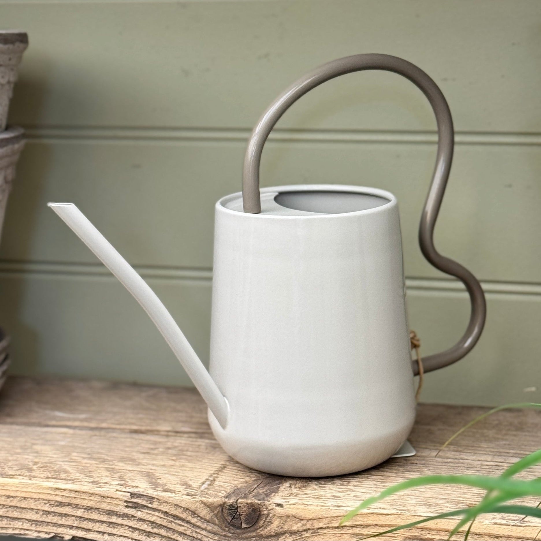 Grey watering can with a curved handle on a wooden surface with potted plants in the background.