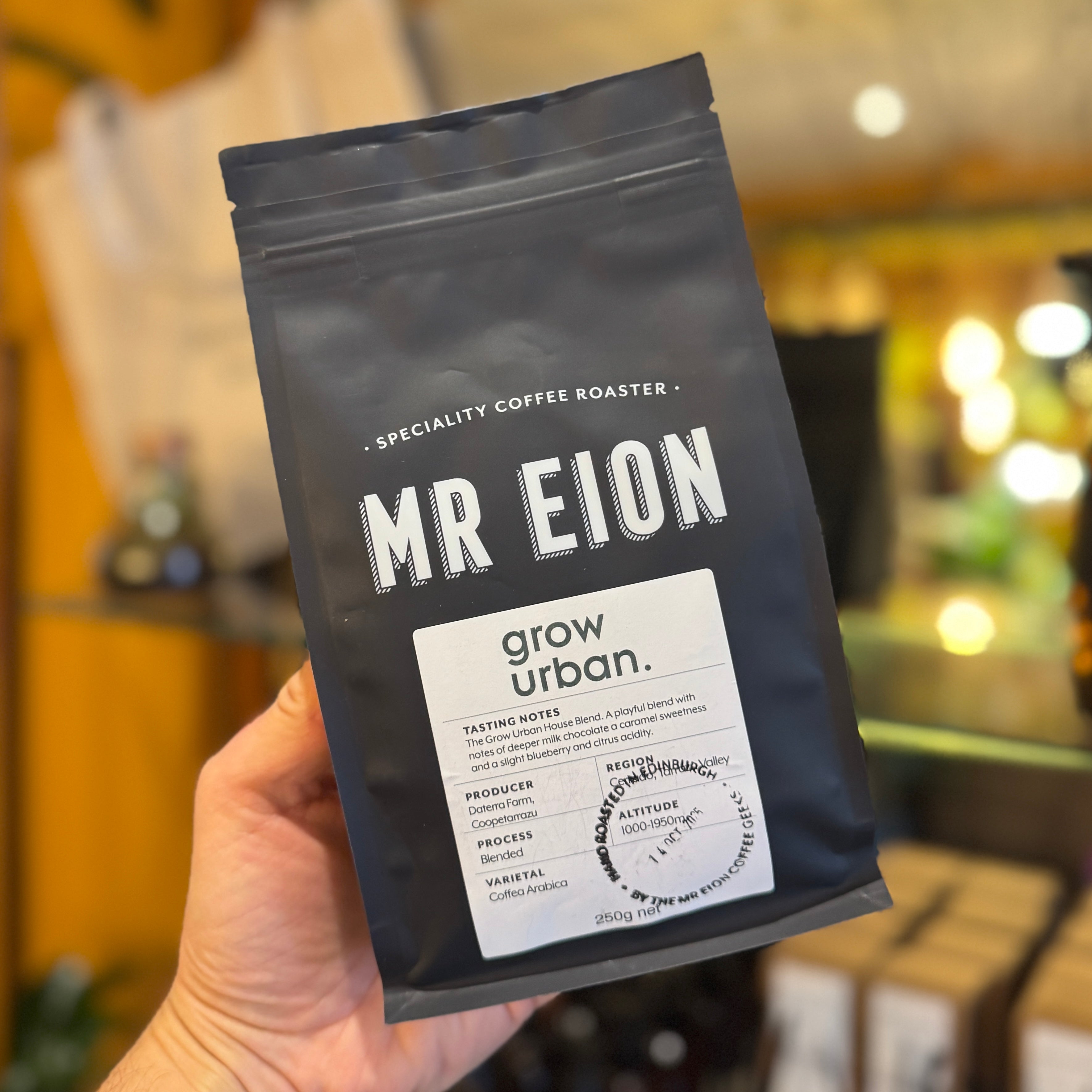 Hand holding a black bag of grow urban coffee beans labeled 'MR EION' with a blurred indoor background.