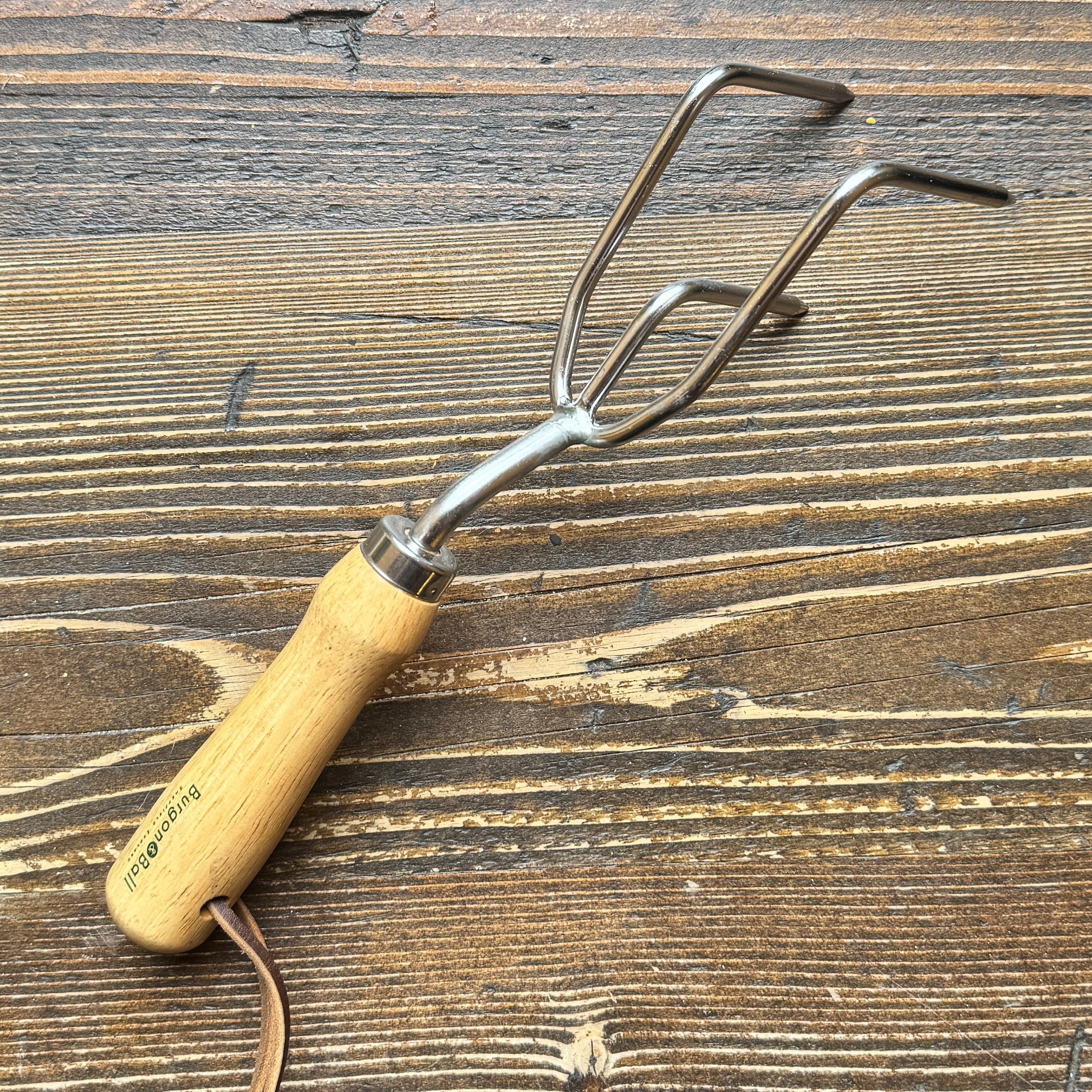 A three-pronged hand fork garden tool with a light wood handle and a brown leather wrist strap, resting on a rustic dark wood background.