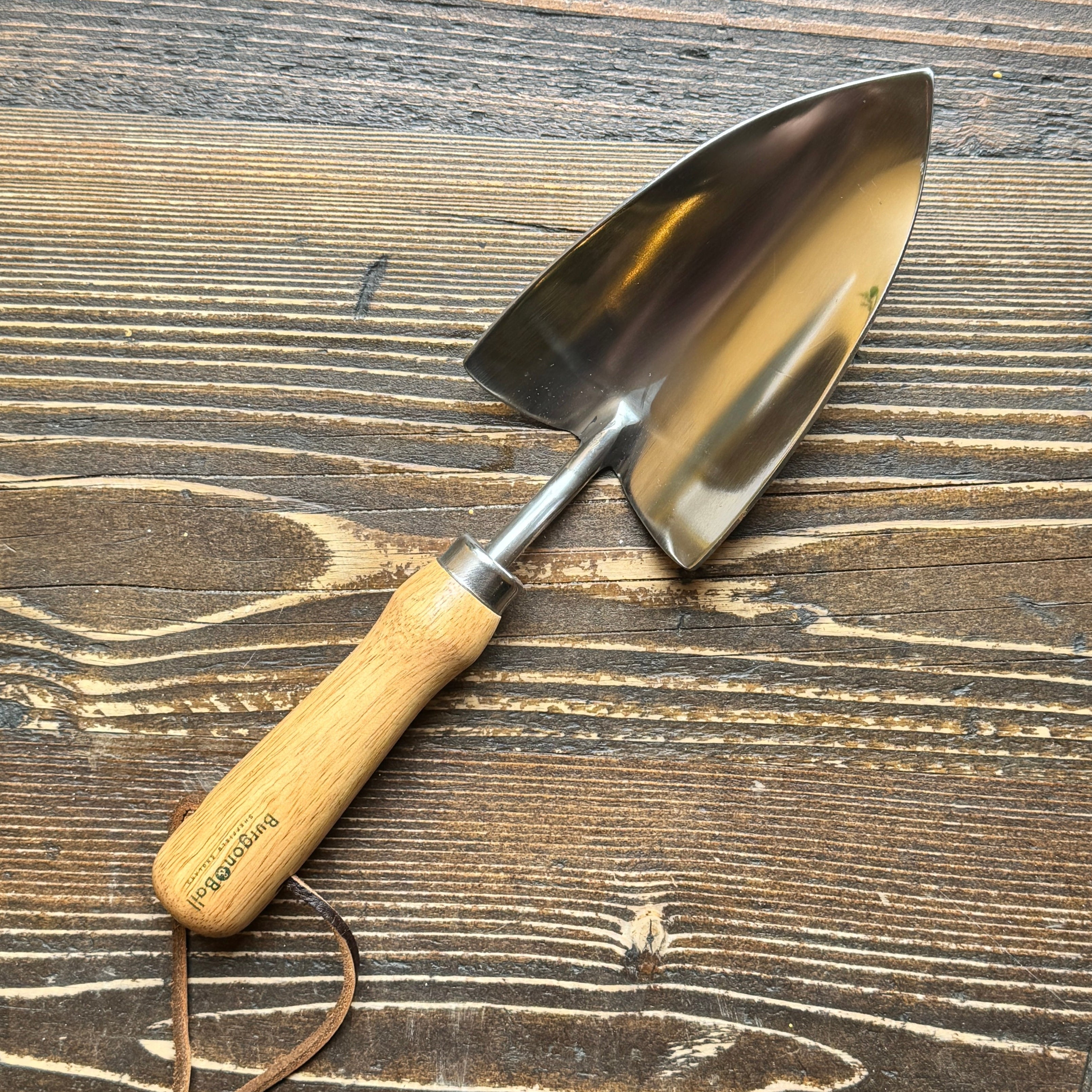 A small hand trowel with a polished steel blade and a light wood handle, resting on a dark brown wooden surface. The handle is printed with the brand name 'Burgon & Ball'