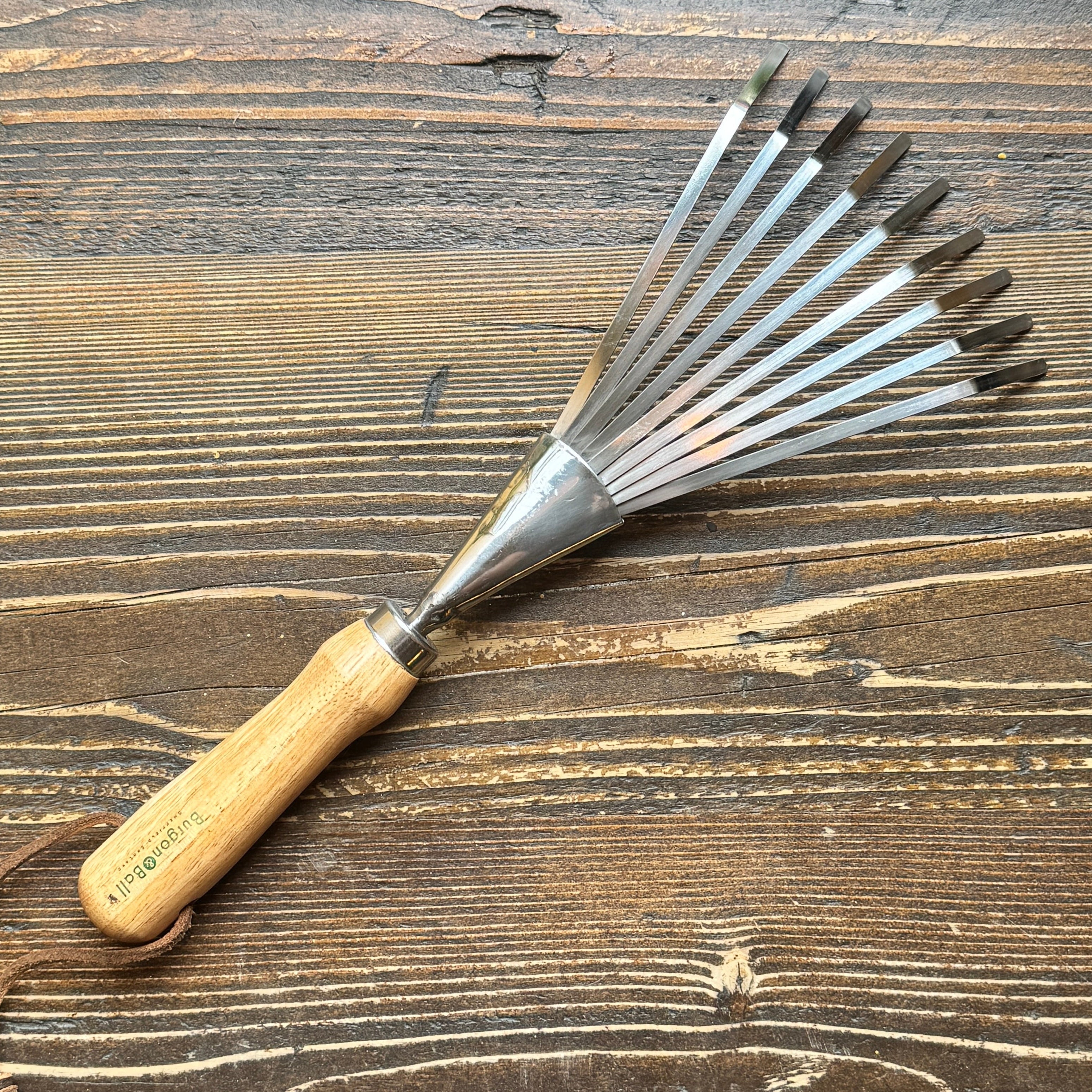 A Shrub Rake with a light wood handle, a silver metal guard, and multiple dark steel blade tips, displayed diagonally on a rustic dark wood background.