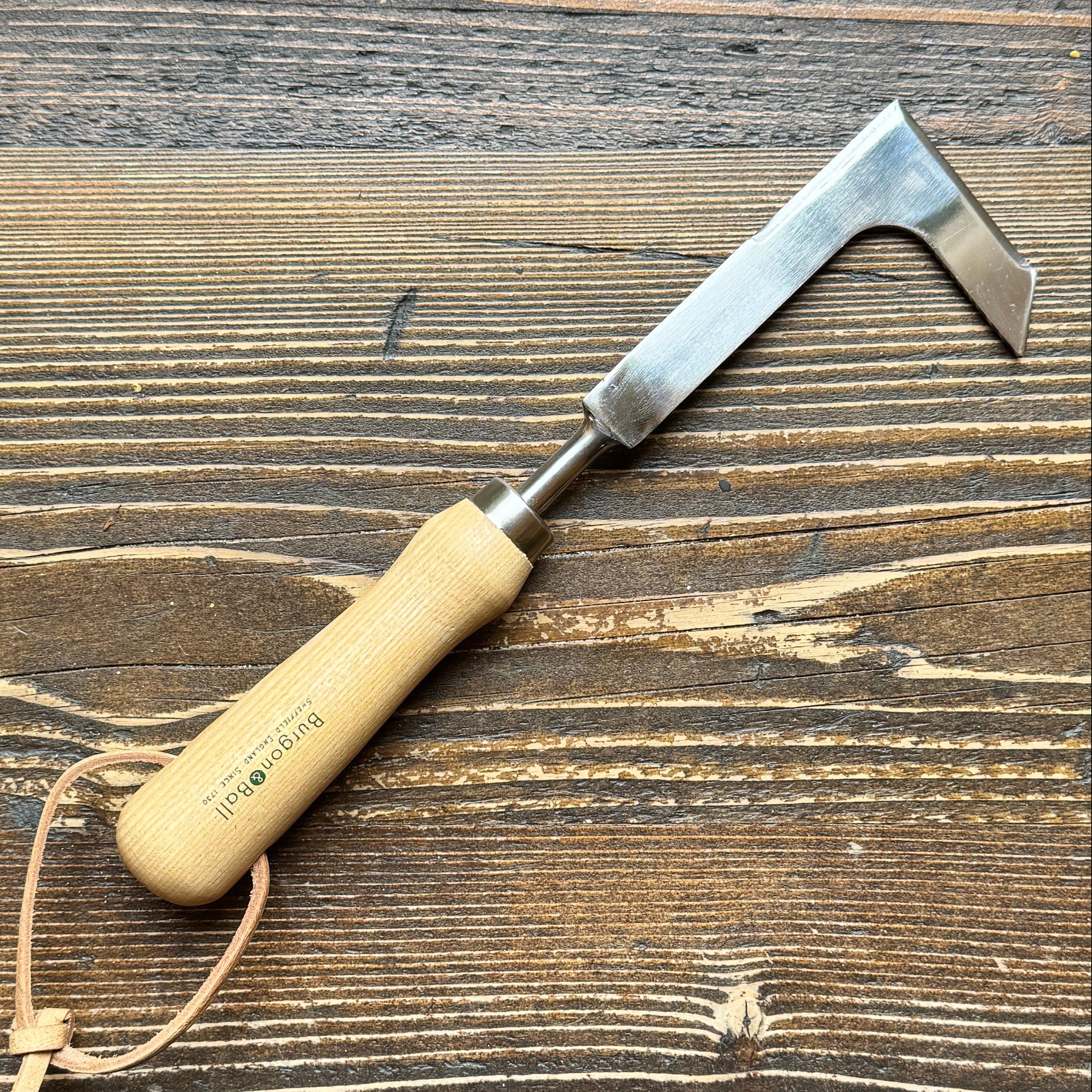 A hand blocking paving knife and weeding tool with a light natural wood handle, a silver metal blade, and a small tan leather loop, placed diagonally on a dark brown wood plank surface. The handle is printed with the brand name 'Burgon & Ball'