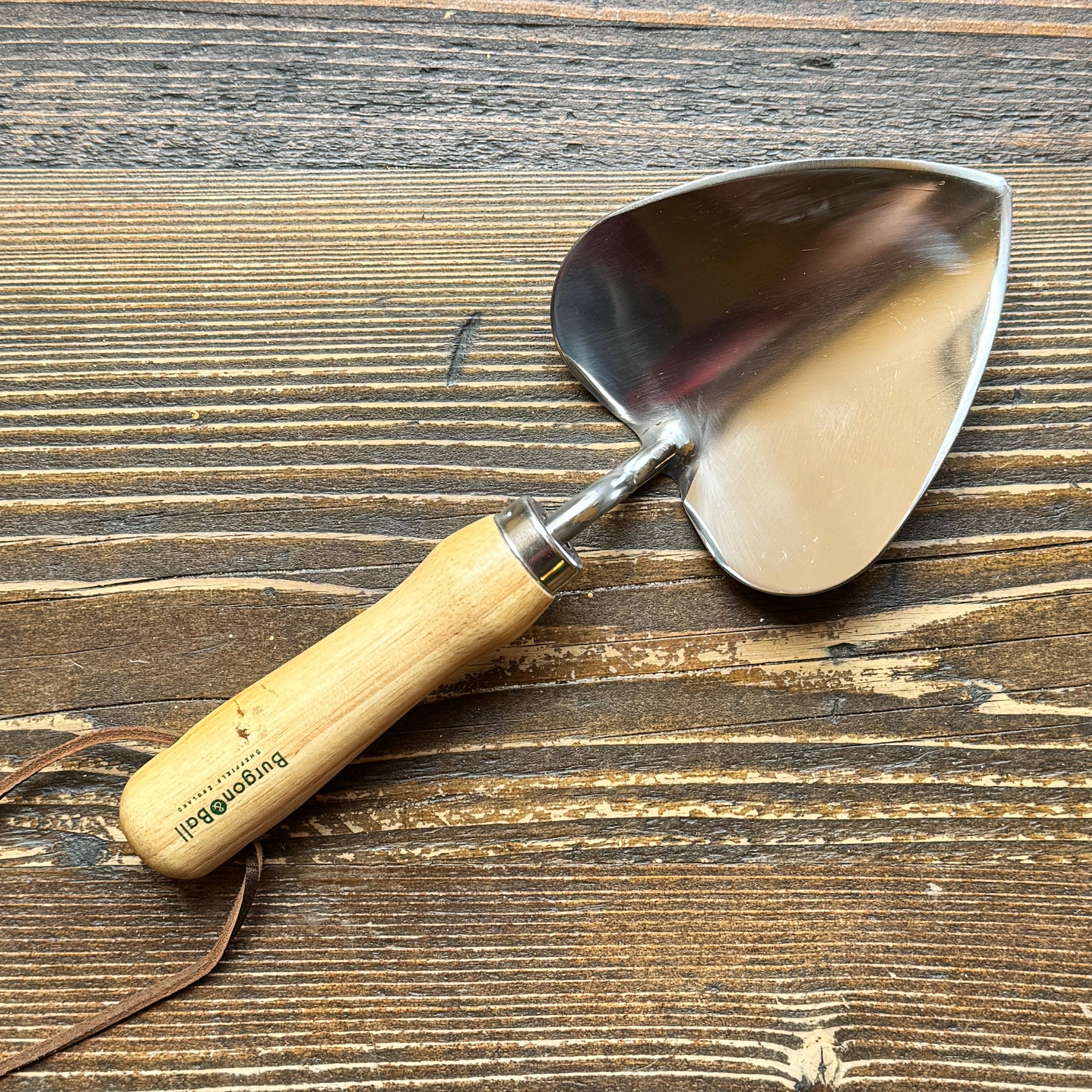 A small heart-shaped hand trowel with a polished steel blade and a light-coloured wooden handle, resting on a rustic dark wood surface. The handle is printed with the brand name 'Burgon & Ball'.