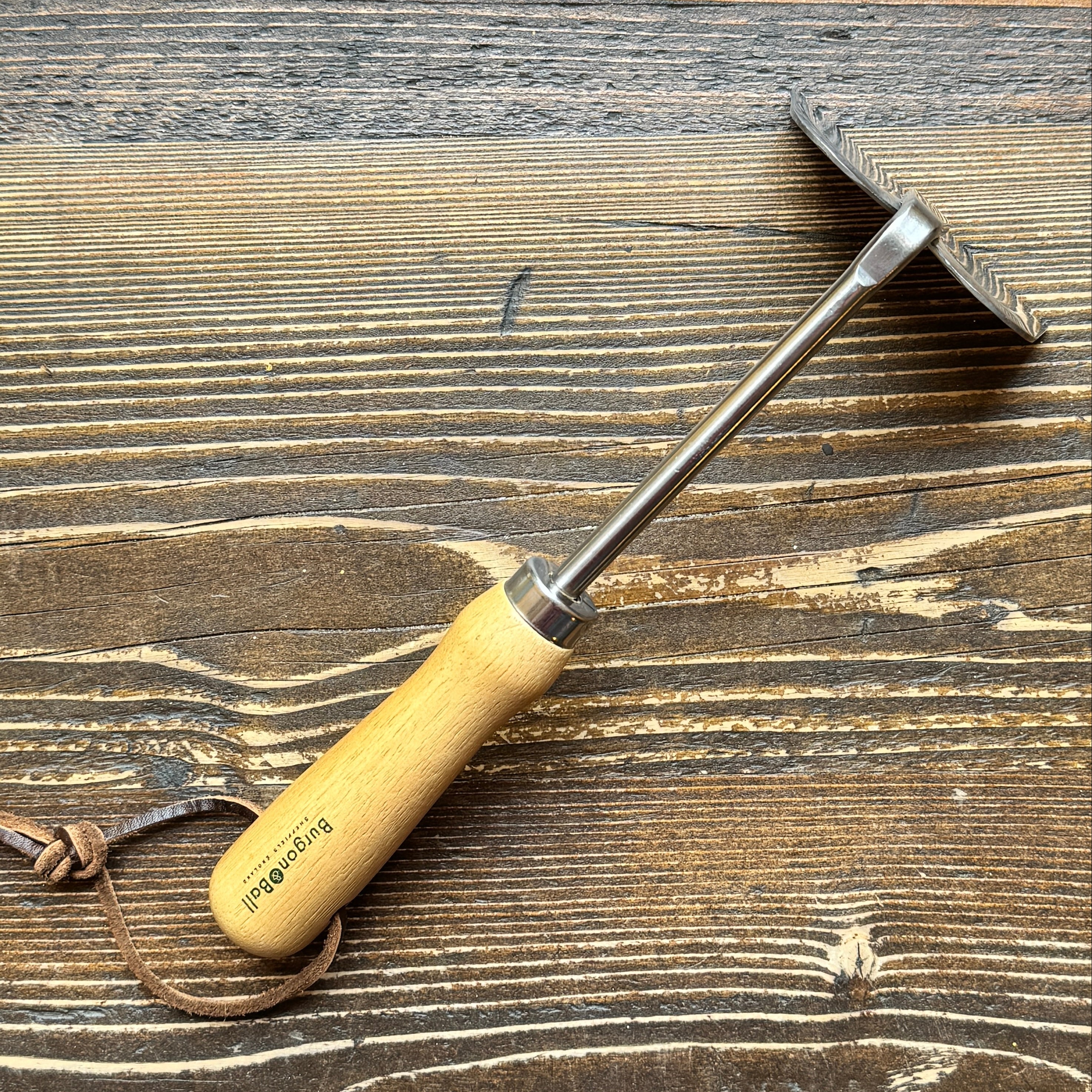 A Hand Hoe with a light wood handle and a silver metal blade, placed diagonally on a dark brown wooden surface. The handle is printed with the brand name 'Burgon & Ball'.