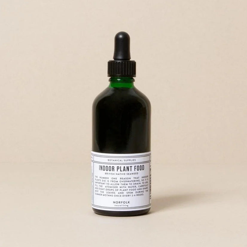 A dark glass dropper bottle labeled 'INDOOR PLANT FOOD' by Norfolk Natural Living, set against a solid beige background.