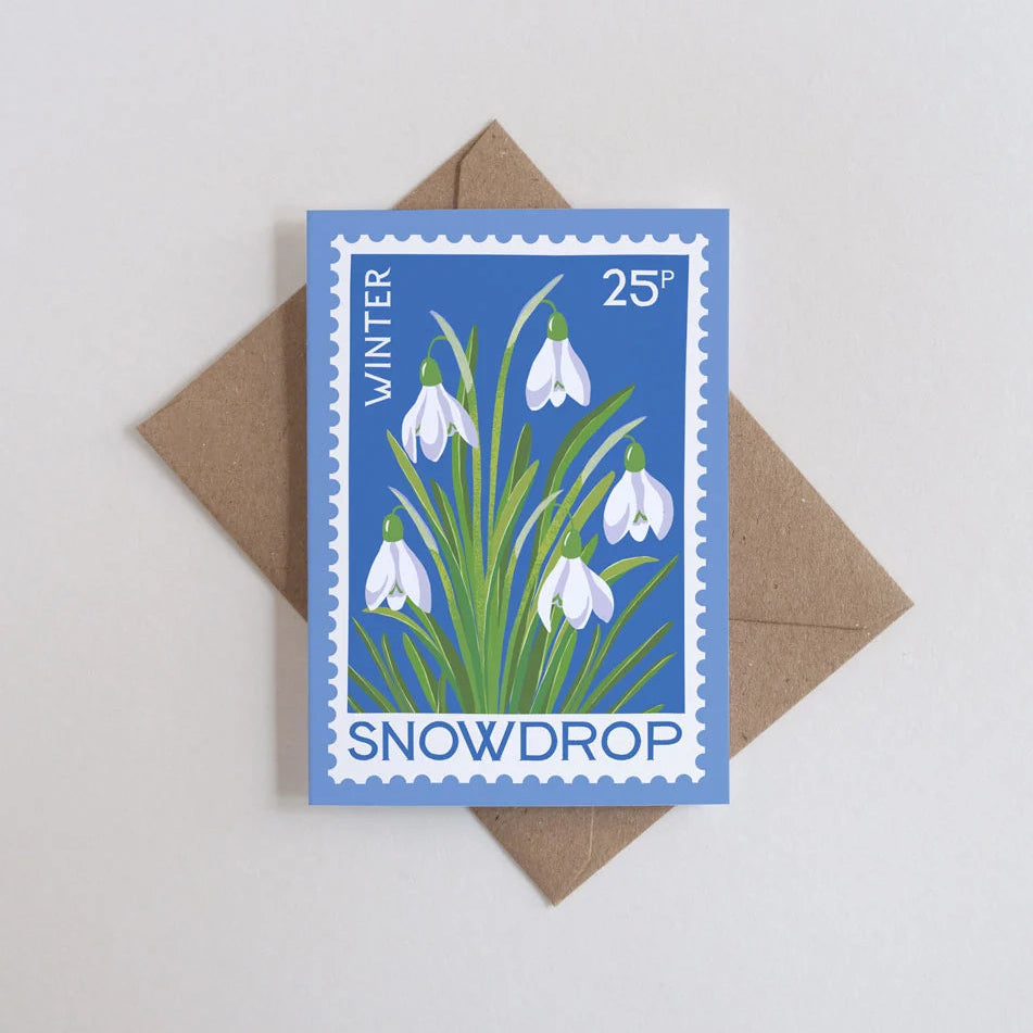 A square greeting card styled like a vintage blue snowdrop postage stamp, placed on top of a brown kraft envelope against a light gray background.