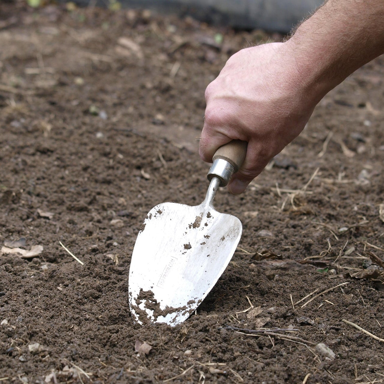 Hand using a Sneeboer Transplanting Trowel to work the soil in a garden
