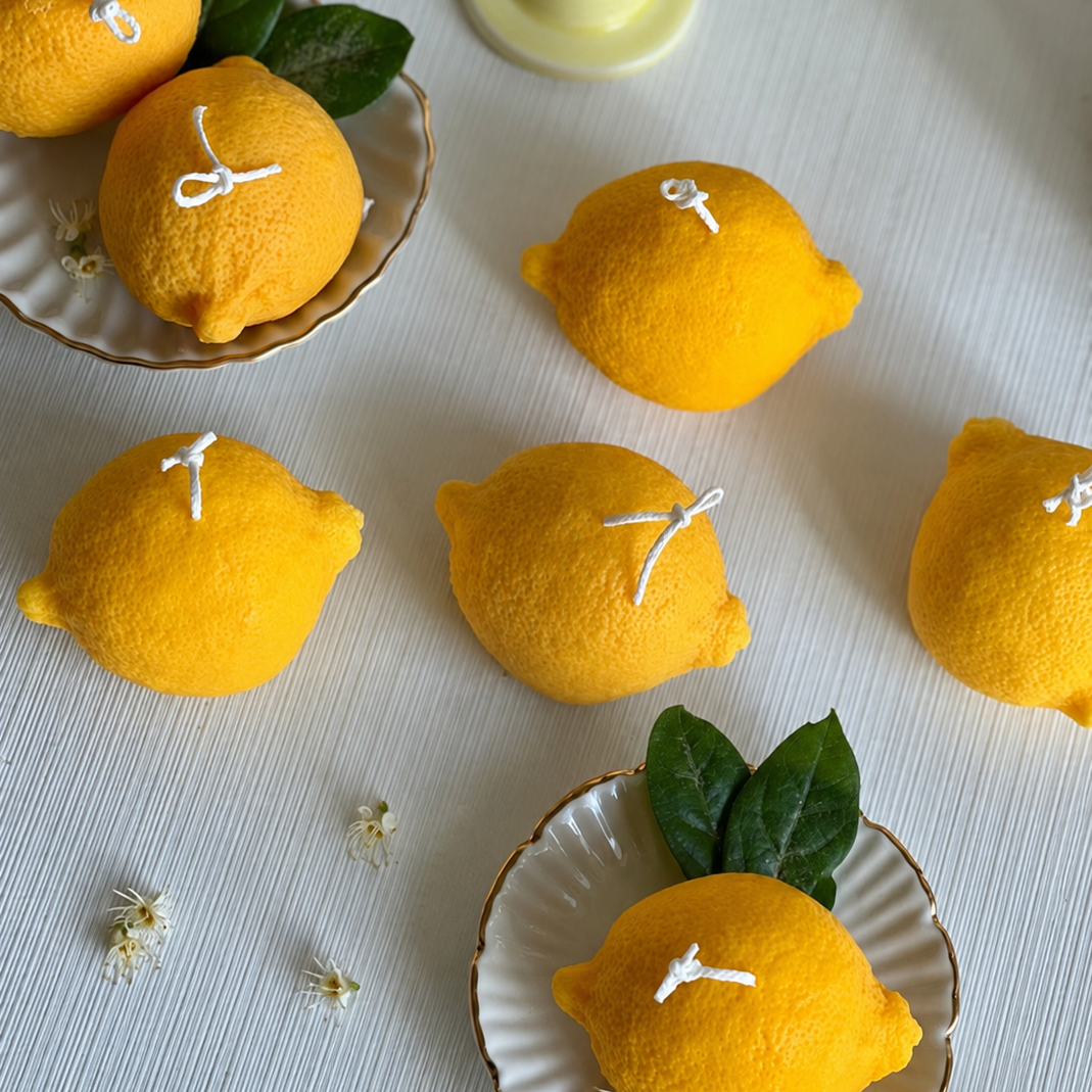 Yellow lemon-shaped candles with green leaves and white wicks, styled on a white textured surface with decorative plates and small flowers.