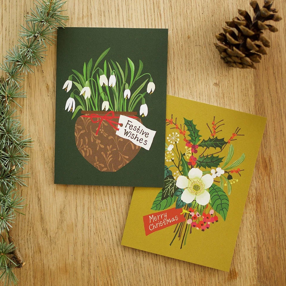 Two illustrated Christmas greeting cards arranged on a wooden surface with a pine branch and a pinecone. One card is dark green with snowdrops and a banner reading 'Festive Wishes,' and the other is mustard yellow with a floral arrangement and a banner reading 'Merry Christmas.