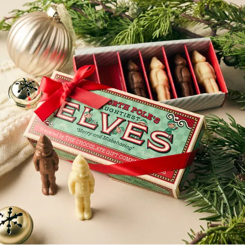A boxed set of Elf chocolate figures and matchbox-style packaging arranged with holiday props on a cream surface. The front box is teal with red lettering and a red ribbon, with an open box behind showing brown and cream elf figures inside.