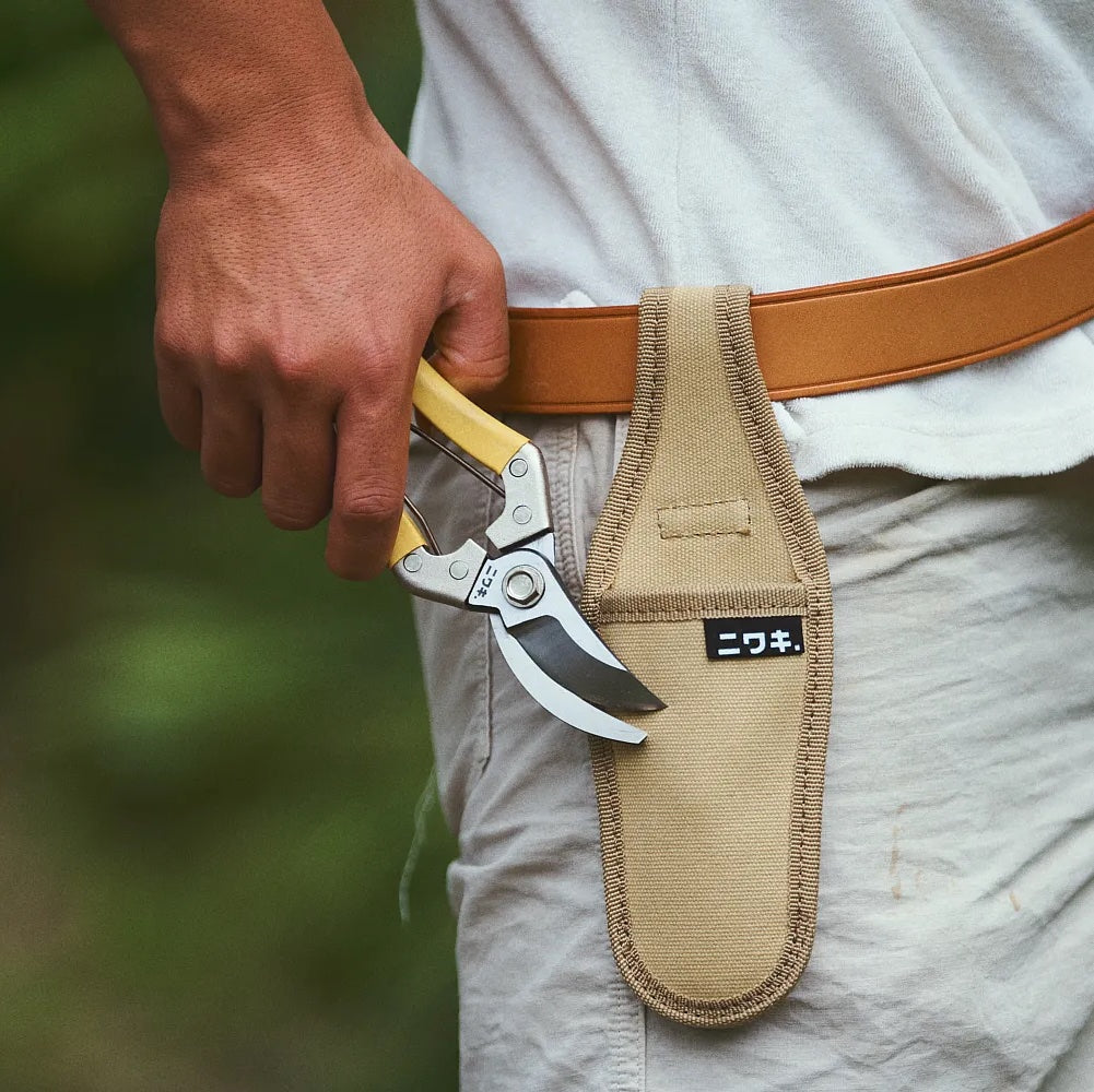 Hand holding a pair of garden shears in a tan sheath with a blurred green background