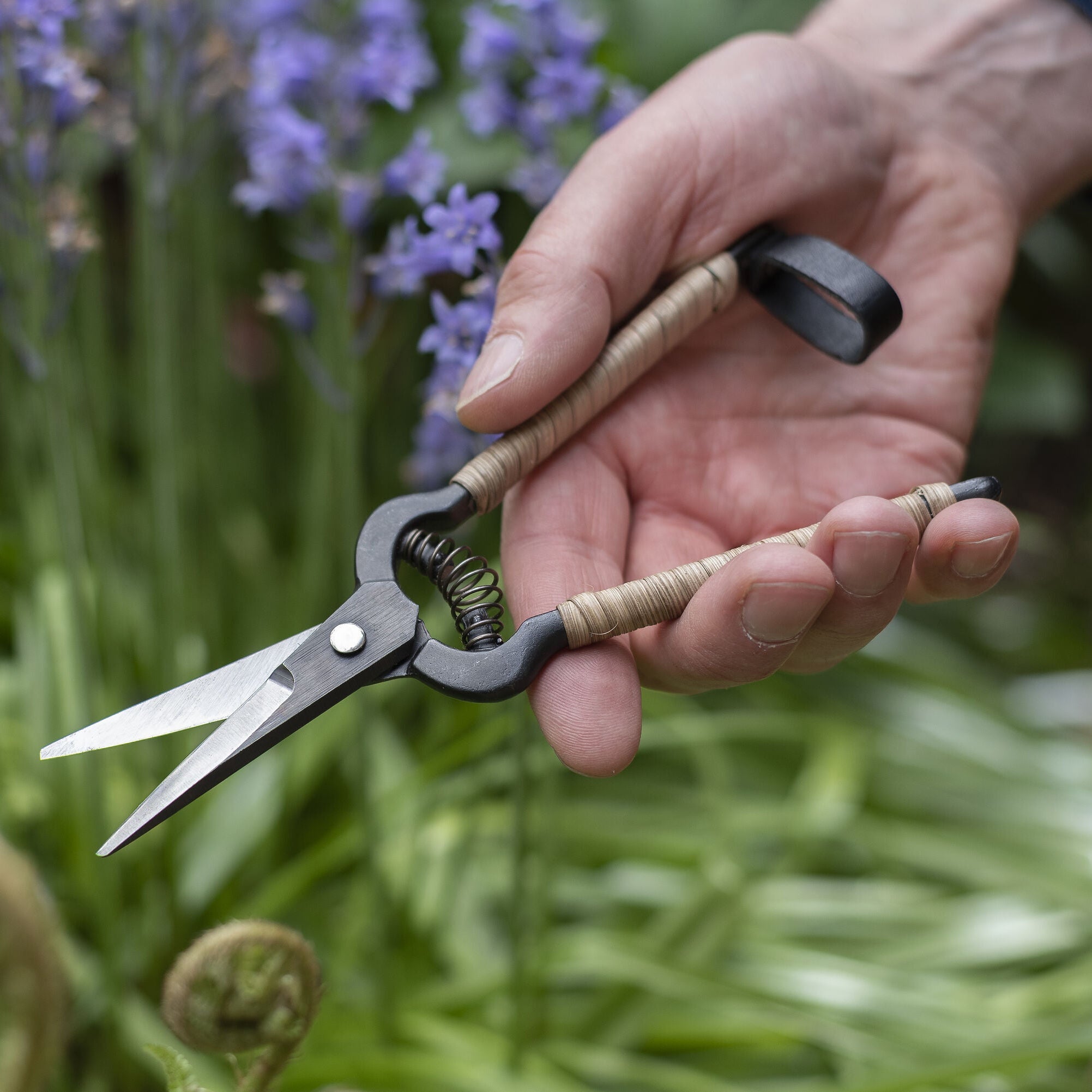 Hand holding Niwaki Rattan Snips with a blurred background of greenery and purple flowers