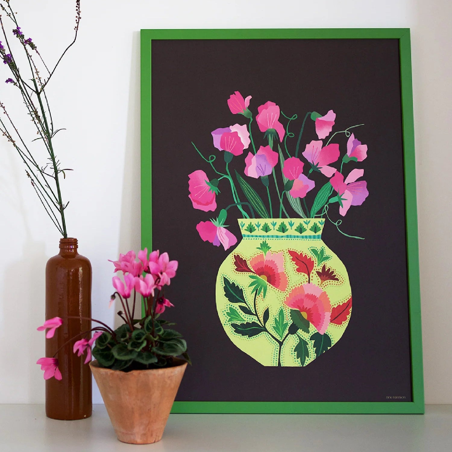 Framed artwork of a floral design with pink flowers and green leaves on a black background, placed on a white surface.